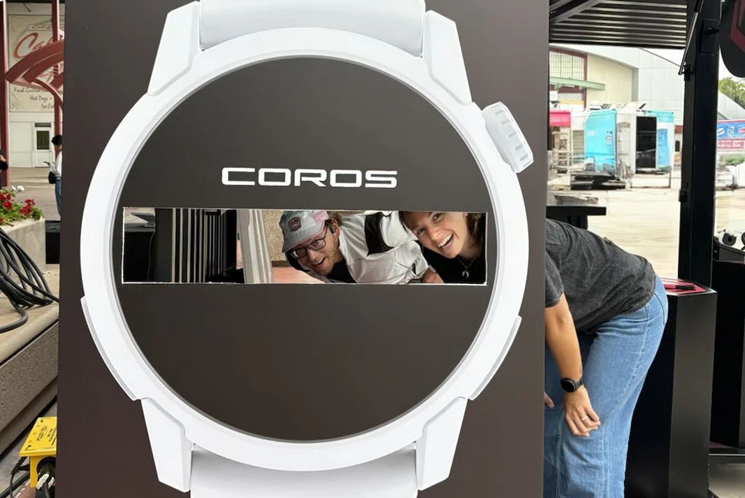 Due membri del team COROS sorridono attraverso un grande espositore a forma di orologio.
