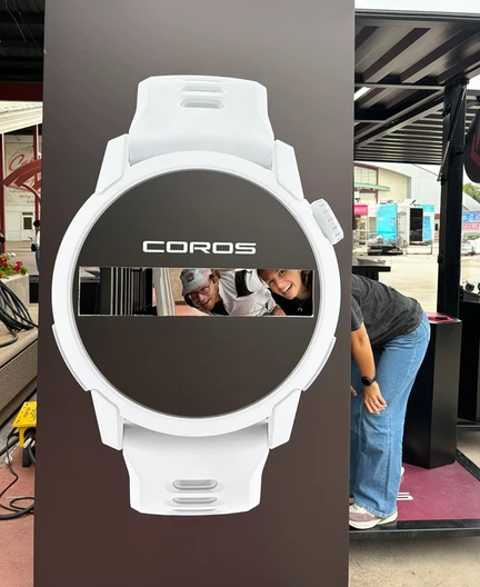 Due membri del team COROS sorridono attraverso un grande espositore a forma di orologio.