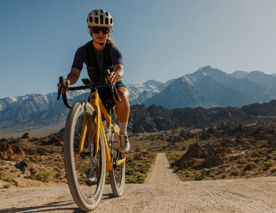 Cyclist riding a yellow bike on a dirt road with mountains in the background.
