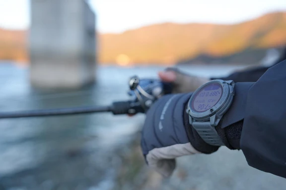 Close-up of a COROS watch on a wrist holding a fishing rod near water.