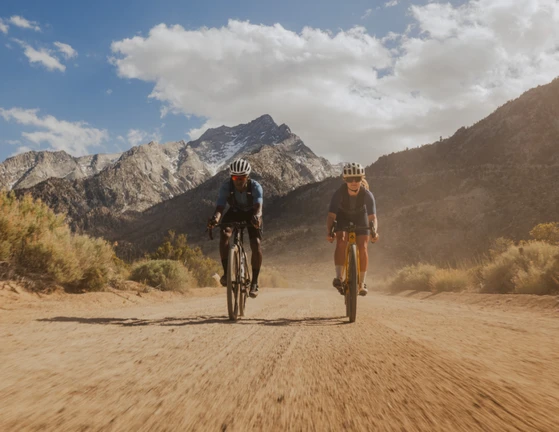 Two cyclists riding on a dirt road with mountains in the background.