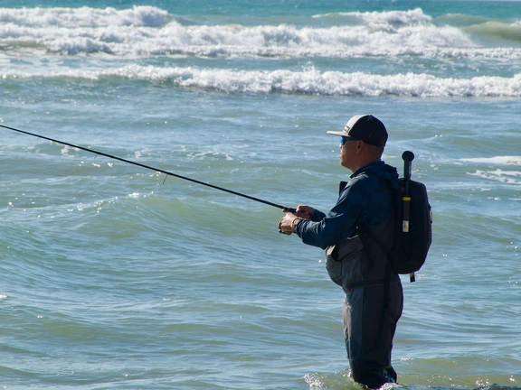 Man in waders surf fishing in the ocean with a backpack.