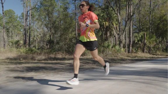 A woman running on a road lined with trees wearing sunglasses.